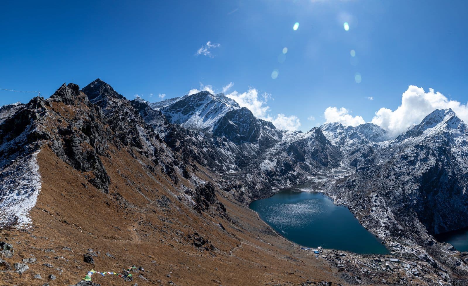 Gosainkunda lake in a rocky bowl below snow peaks, with a trail traversing the brown hillside and small stone lodges on the far shore
