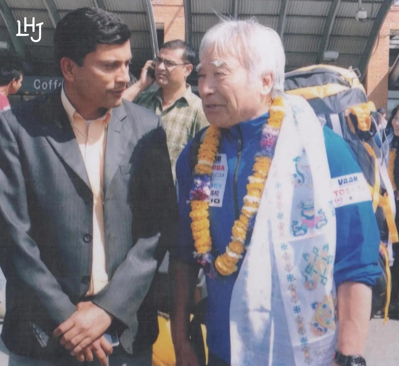 Yūichirō Miura, garlanded with marigolds and wearing a white ceremonial scarf over a blue expedition jacket, in conversation with Deepak Rimal in a grey suit at Tribhuvan International Airport.