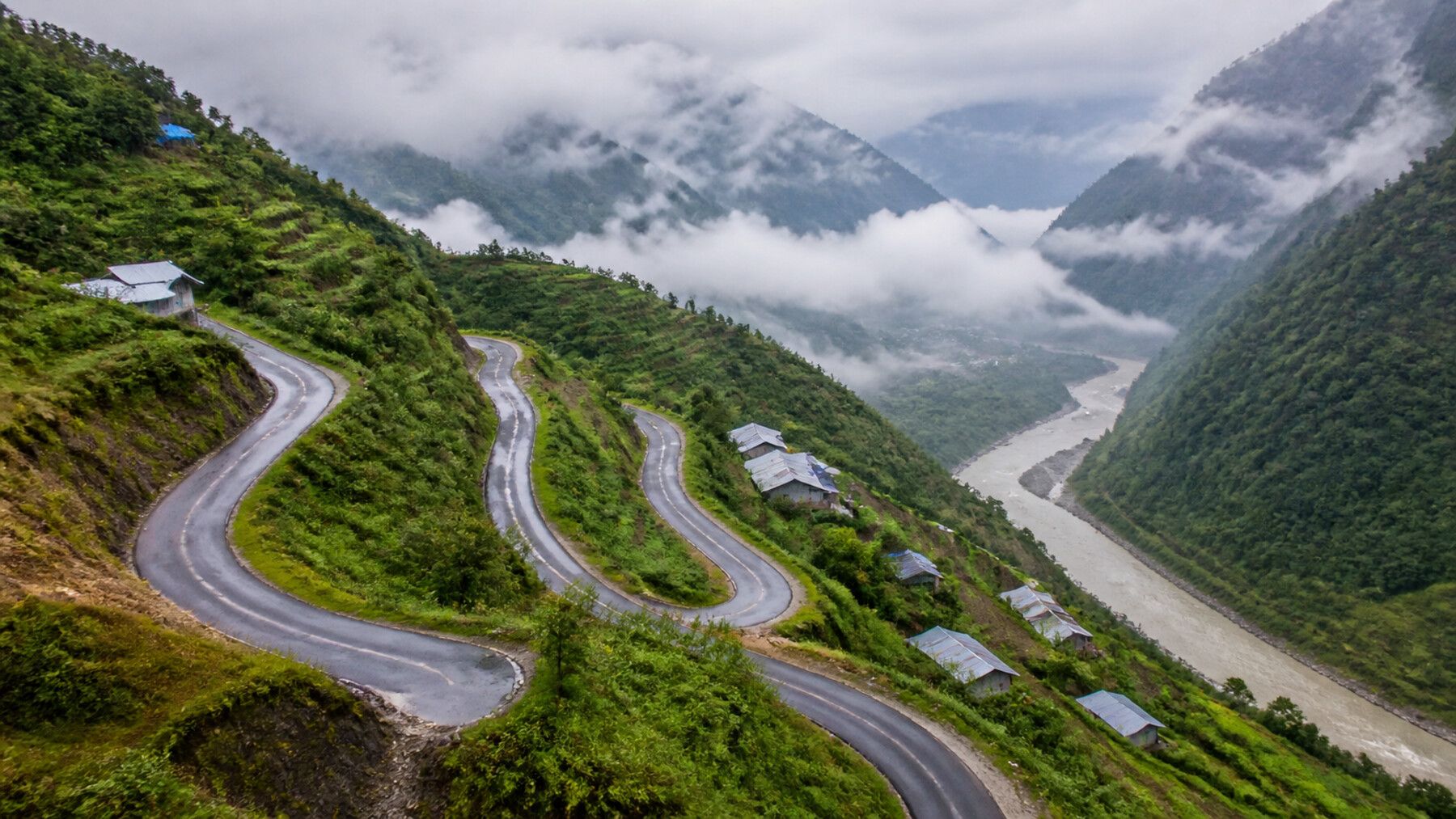 A narrow mountain road switchbacking down a steep terraced hillside into a cloud-filled river valley, with small tin-roofed houses scattered along it.