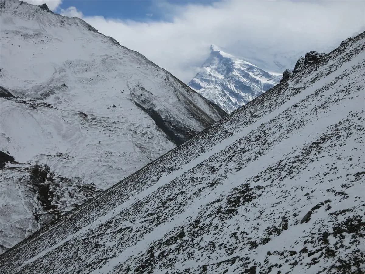 Snow covers the steep sides of a mountain valley beneath broken cloud near Thorong La.