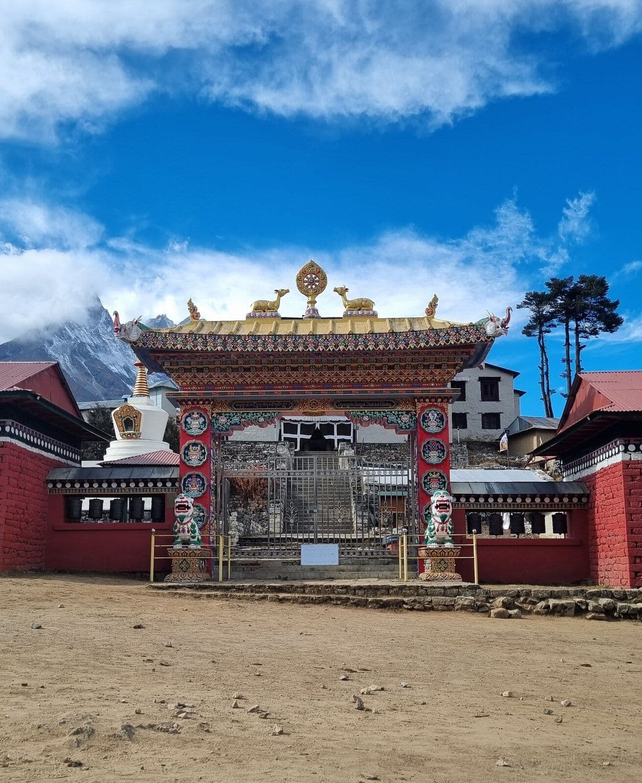 The decorated entrance gate of Tengboche Monastery beneath a blue sky, with a stupa and mountain ridge behind it.