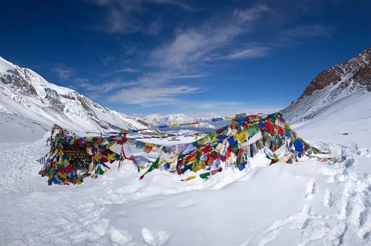 Prayer flags stretch across a snow-covered hollow at Thorong La beneath a blue sky.