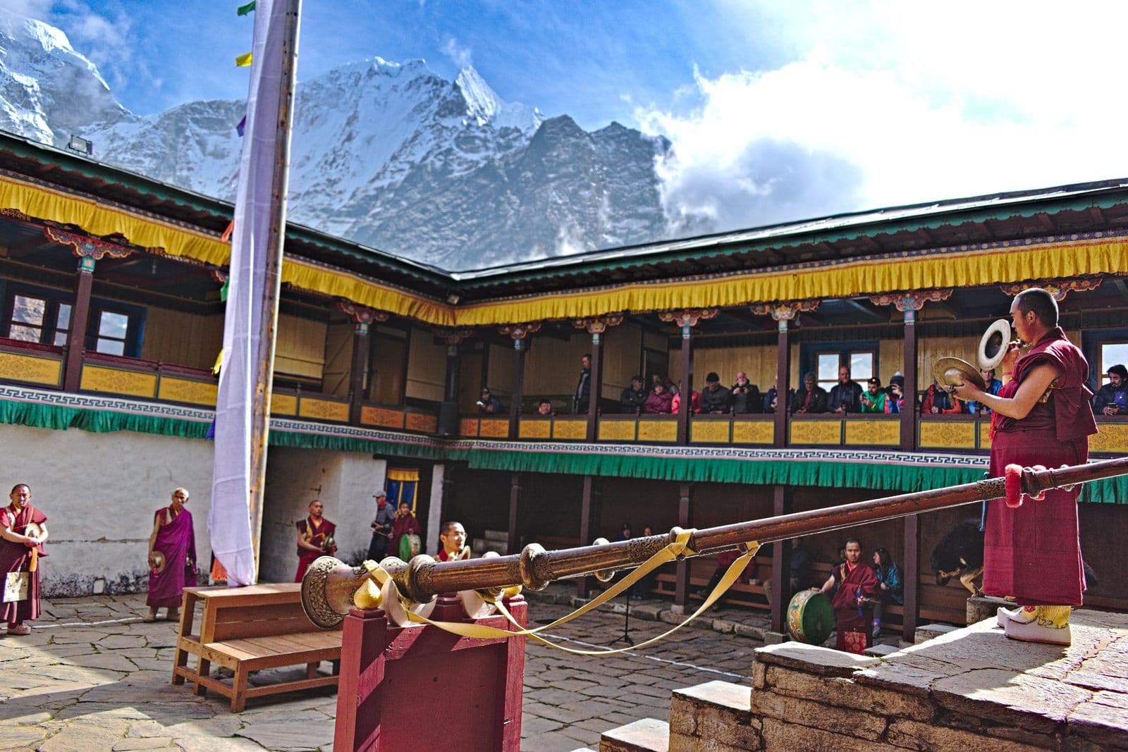 Monks with cymbals and a long dungchen horn in the Tengboche courtyard during Mani Rimdu, with trekkers watching from the upper gallery and snow peaks above the roofline
