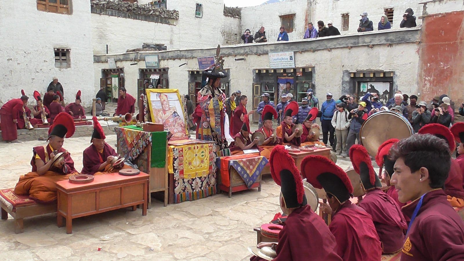 Monks in red robes and black crested hats playing cymbals and drums around a costumed dancer in the Lo Manthang courtyard, with visitors watching from the rooftops