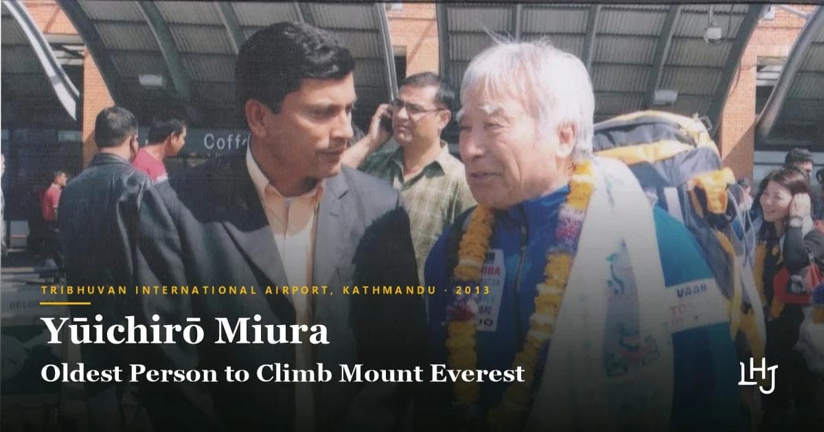 Yūichirō Miura, wearing marigold garlands and a white ceremonial scarf over a blue expedition jacket, in conversation with Deepak Rimal in a grey suit at an airport arrivals area. Overlaid text reads: Tribhuvan International Airport, Kathmandu, 2013. Yūichirō Miura, Oldest Person to Climb Mount Everest.