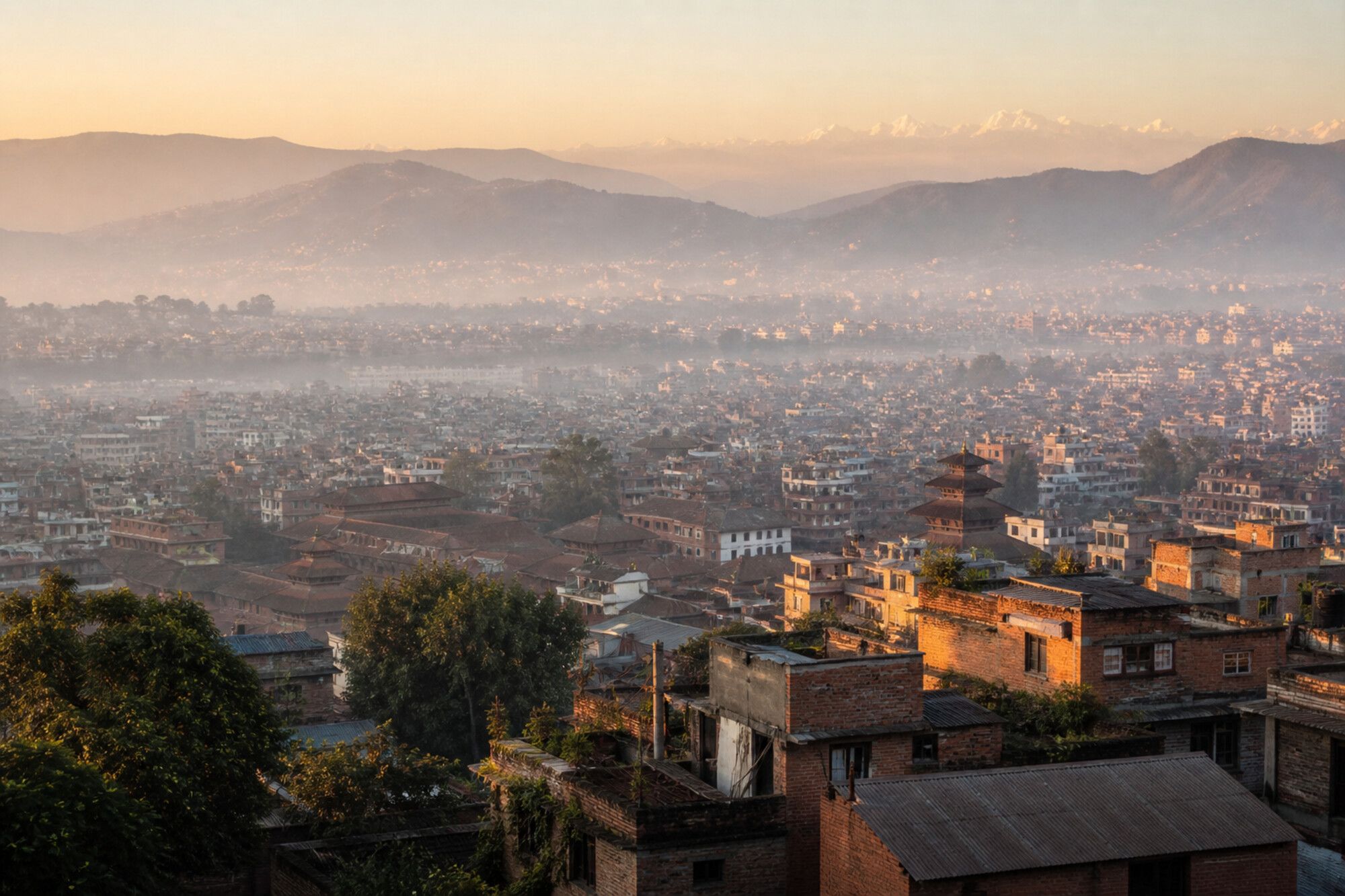 A hazy early-morning view across the Kathmandu Valley, with brick rooftops and tiered temple roofs in the middle distance, terraced green hills behind, and a faint line of snow peaks on the horizon.