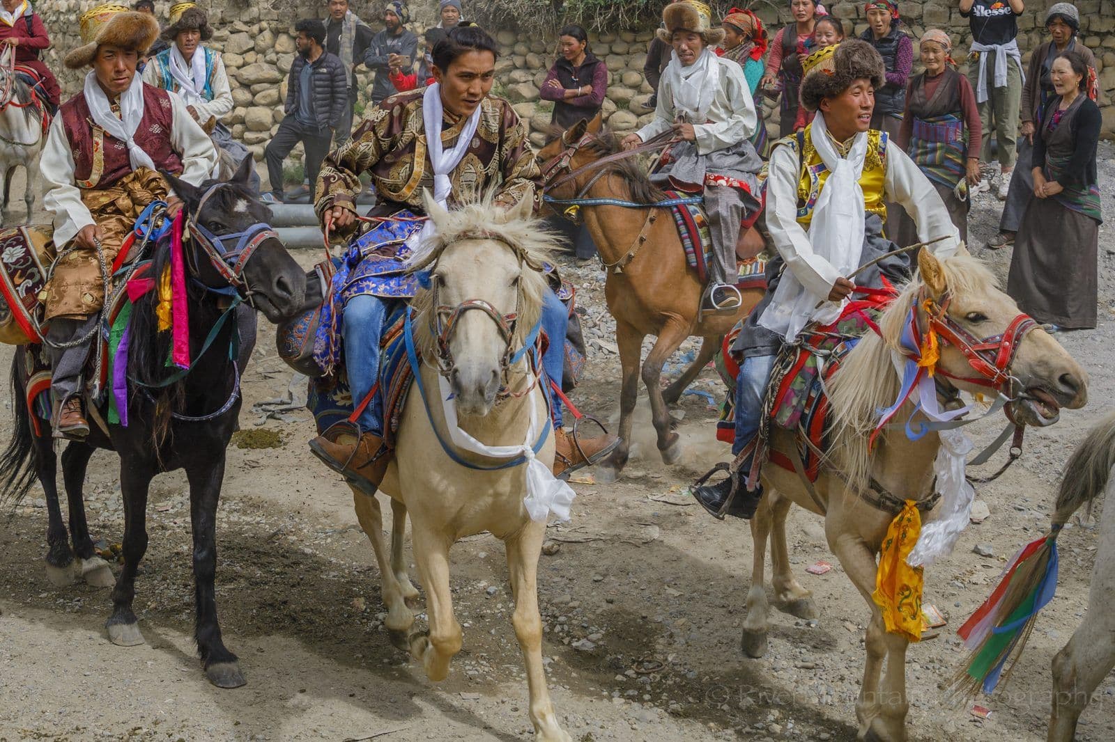 Four riders in fur-brimmed hats and brocade robes on horses decorated with coloured ribbons at Yartung, with village spectators watching from behind a stone wall
